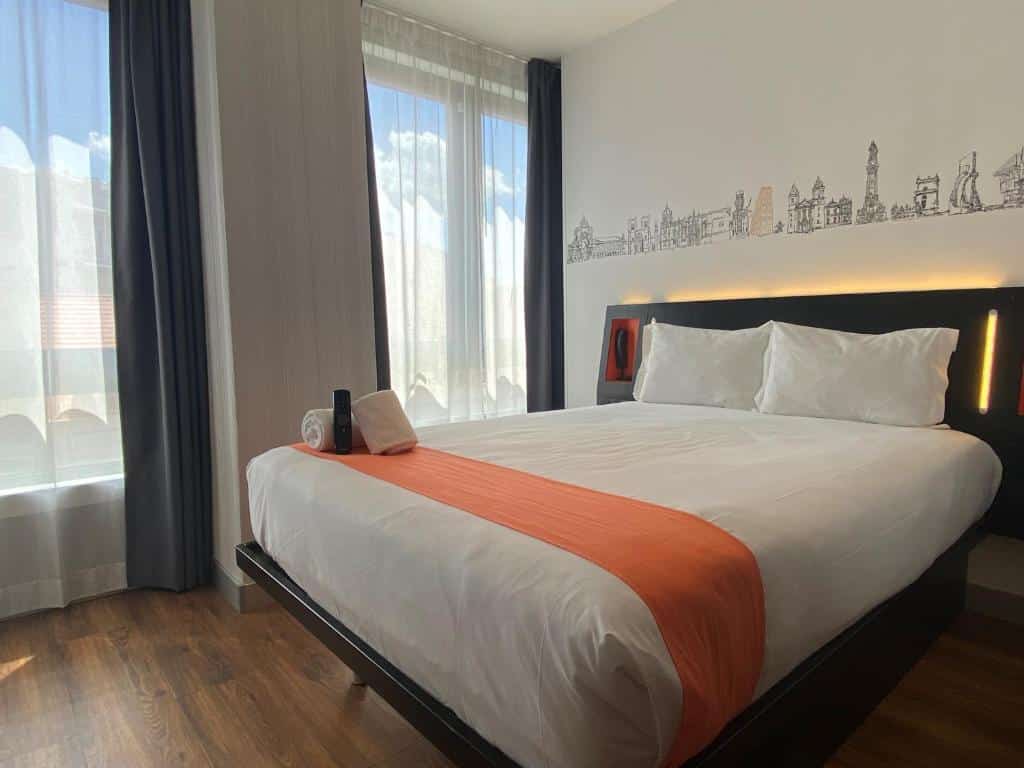 Quarto do easyHotel Lisbon com uma cama de casal, do lado esquerdo, há duas janelas com cortinas brancas e o chão do ambiente imita madeira.