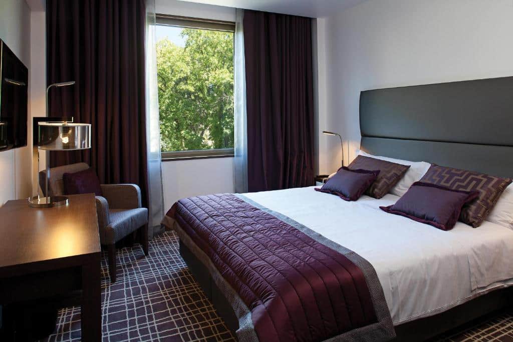 Quarto do NEYA Lisboa Hotel com uma cama de casal, uma janela com cortinas, chão de carpete em tons de branco e roxo, de frente com a cama há uma mesa de madeira com uma televisão sob ela, além de uma poltrona perto da janela