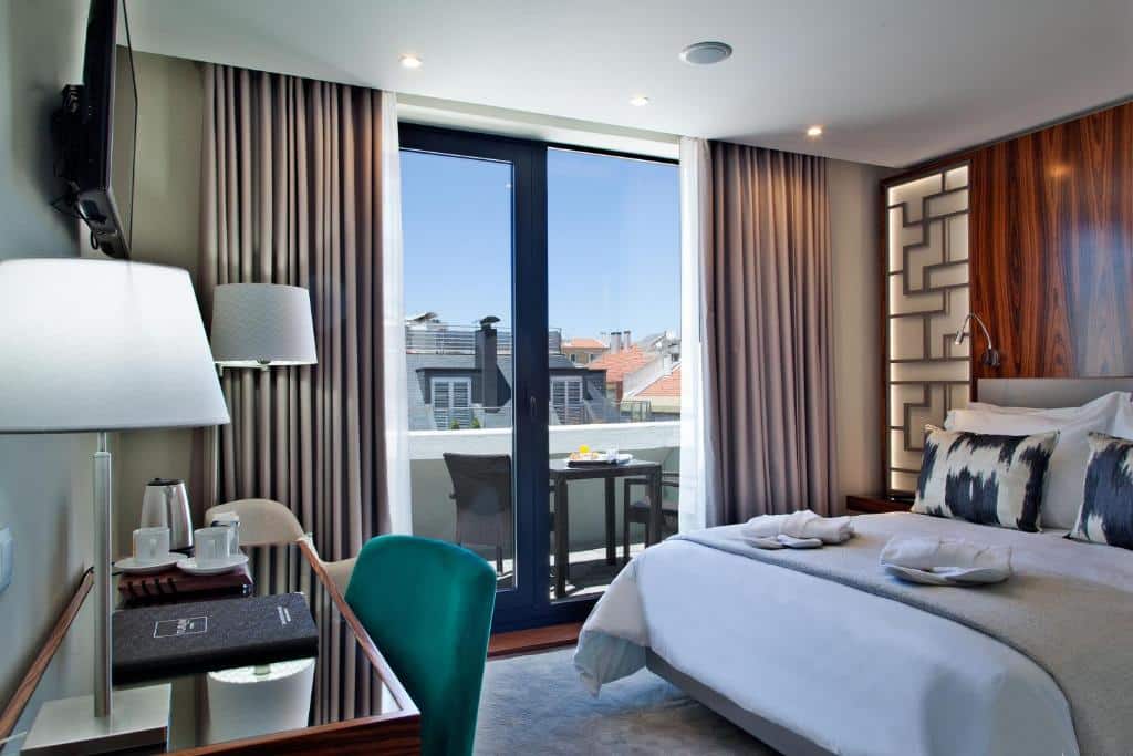 Quarto do TURIM Saldanha Hotel com uma cama de casal com almofadas e toalhas sob ela, do lado esquerdo da cama há uma sacada com cortinas e uma pequena mesa com duas cadeiras, do lado de dentro, de frente para a cama, há uma televisão e uma mesa de escritório com uma poltrona