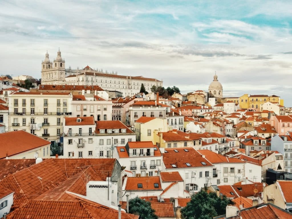Um monte de casinhas de telhado laranja pelas ruas criando uma paisagem bem singular e bonita, no alto, há uma construção antiga, em Lisboa.