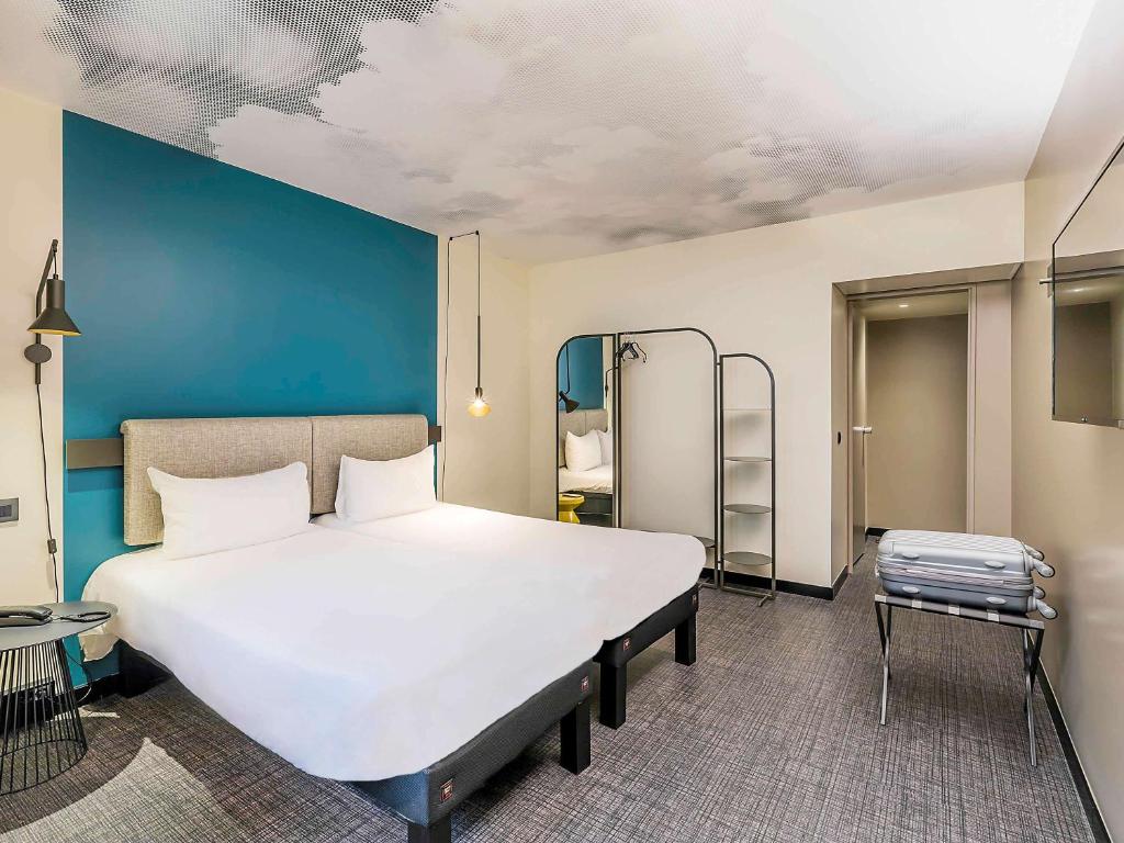 Quarto do Ibis Lisboa Saldanha com uma cama de casal, um espelho de corpo inteiro junto com um cabideiro, há mesinhas de cabeceira com luminárias e uma televisão.
