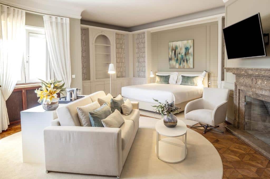 Quarto espaçoso do Luzeiros Suites com uma cama de casal, um pequeno sofá com dois lugares e diversas almofadas sob um apete e de frente para uma televisão, e do esquerdo há uma janela com cortinas e um móvel com utensílios para preparar um café.