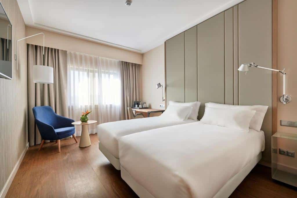 Quarto do NH Lisboa Campo Grande com duas camas de solteiro, uma janela ampla com cortinas cinzas, embaixo da janela há uma pequena mesa com utensílios para fazer café e uma poltrona estofada, na parede de frente para a cama há uma televisão, para representar hotéis baratos em Lisboa.