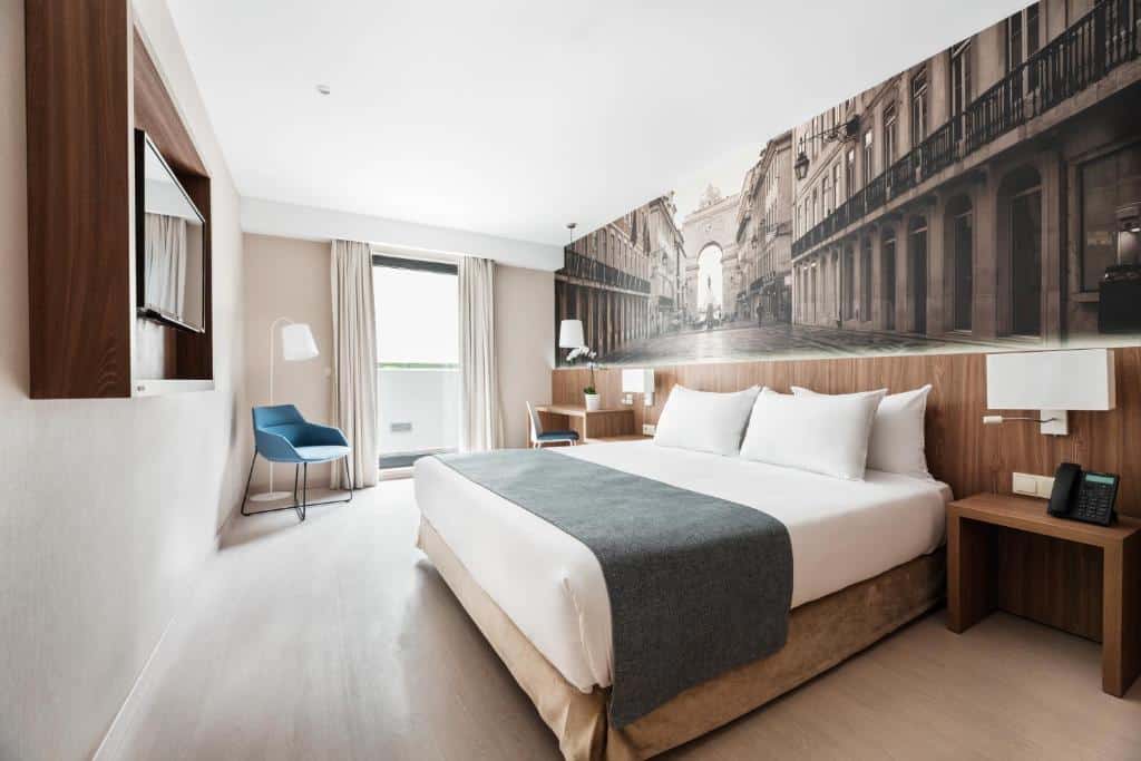 Quarto do Exe Saldanha com uma pequena varanda cm cortinas, uma cama de casal com travesseiros, há também uma mesa de escritório do lado esquerdo da cama, e há uma televisão na parede de frente para a cama