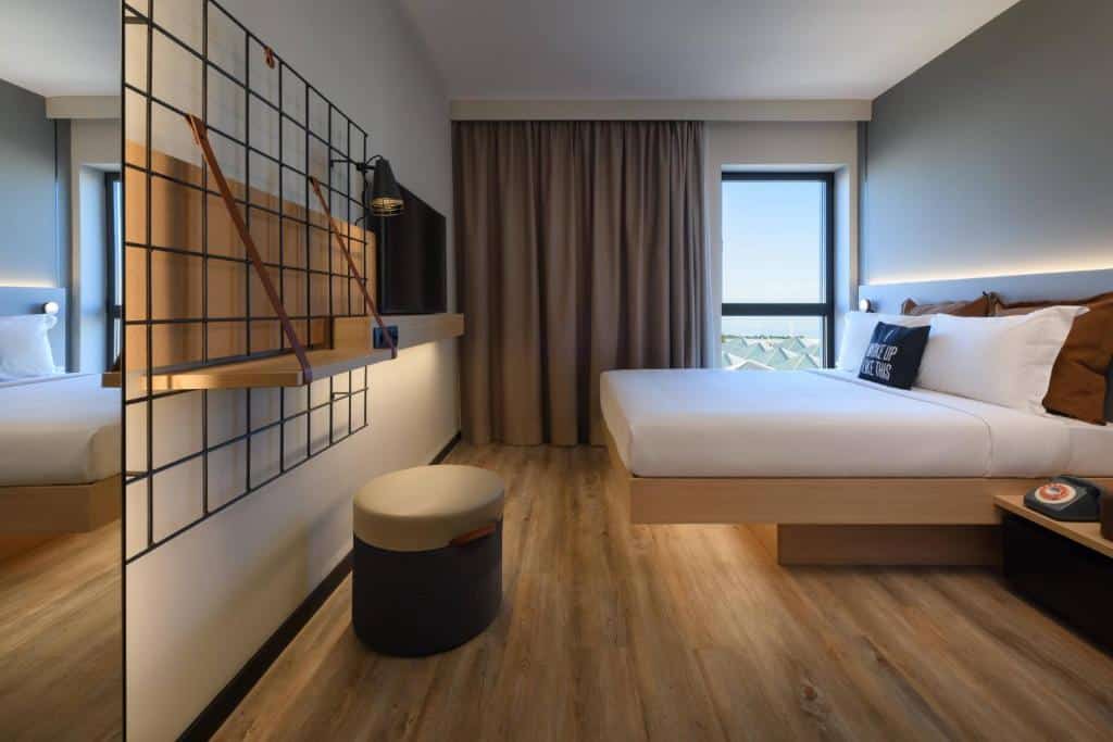 Quarto do Moxy Lisboa Oriente com uma janela com cortinas, uma cama de casal com almofadas e travesseiros, há uma televisão de frente para a cama e, ao lado dela, há uma pequena estante e um bufê, para representar hotéis baratos em Lisboa