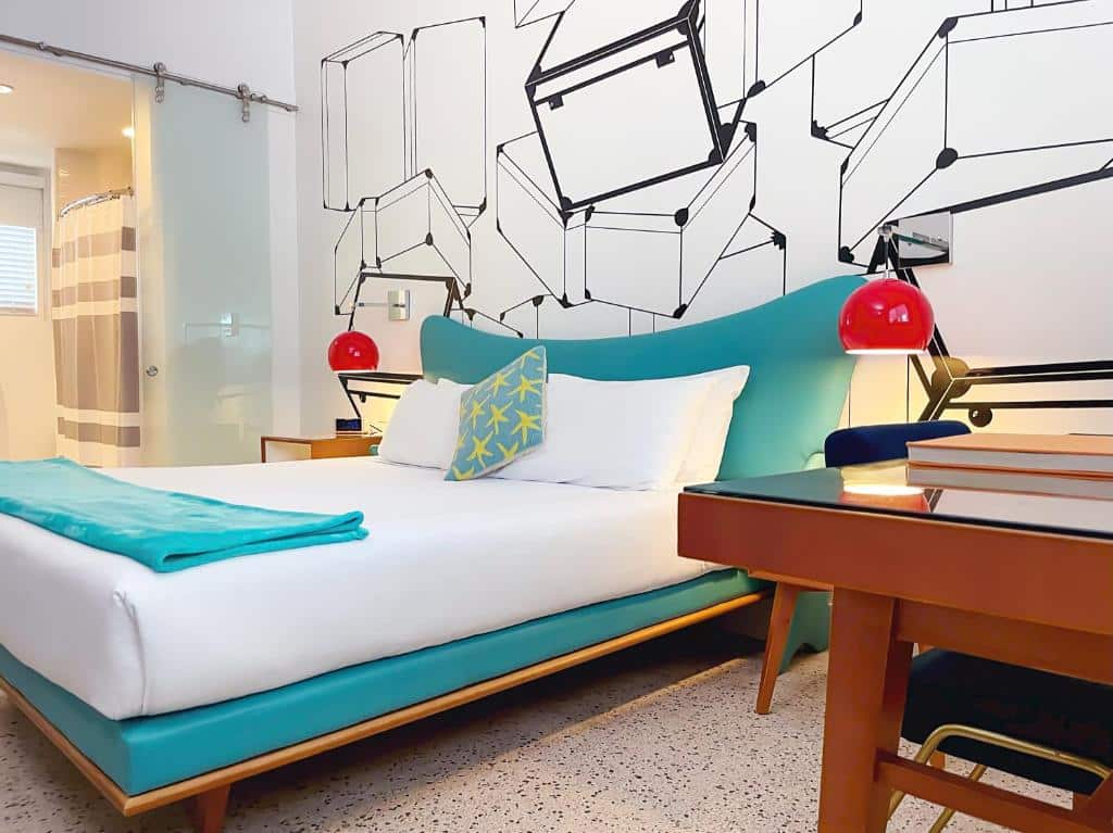 Quarto do The Vagabond Hotel com mesa do lado direito a frente com cadeira ao fundo uma cama de casal no centro do quarto e do lado da cama uma como com luminária, para representar hotéis baratos em Miami.