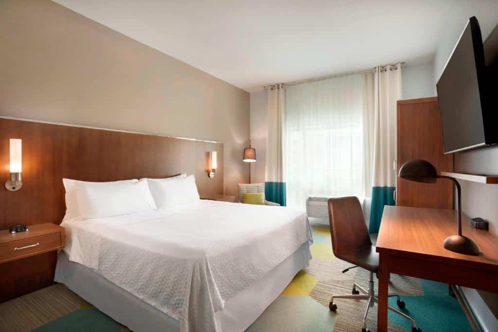 Quarto do Four Points by Sheraton Miami Airport com cama de casal do lado esquerdo da imagem do lado esquerdo da cama uma cadeira em frente a cama uma mesa de trabalho com cadeira e na parede uma TV presa do lado esquerdo da mesa um guarda-roupa.