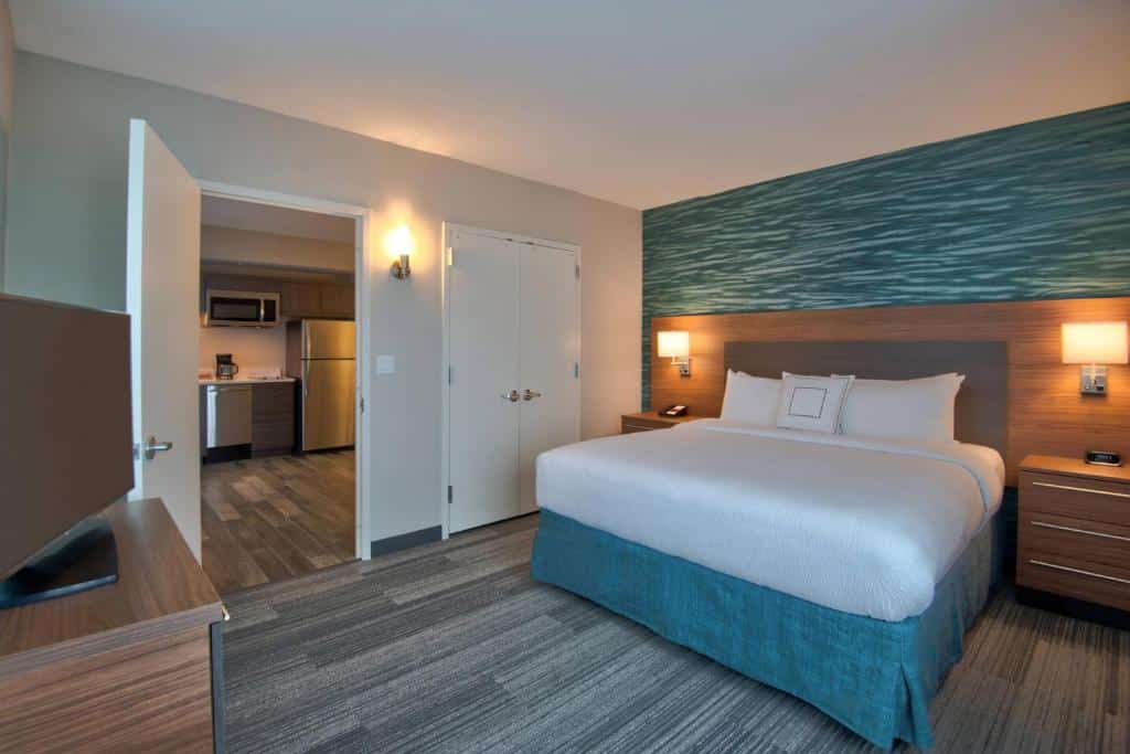 Quarto do TownePlace Suites by Marriott Miami Airport com cama de casal do lado direito da imagem em cada lado da cama uma cômoda com luminária em frente a cama uma cômoda com TV em cima do lado esquerdo da cômoda uma porta que dá acesso a uma cozinha compacta, para representar hotéis baratos em Miami.