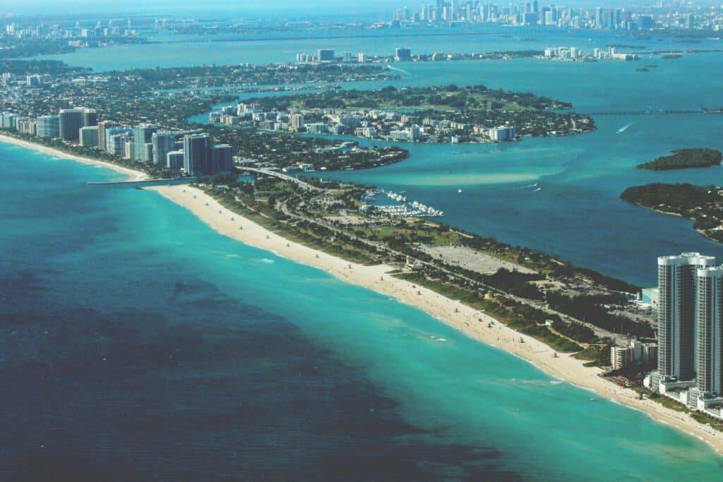 Imagem da praia Miami durante o dia com mar azul do lado esquerdo da imagem e do lado direito faixa de areia com prédios. 