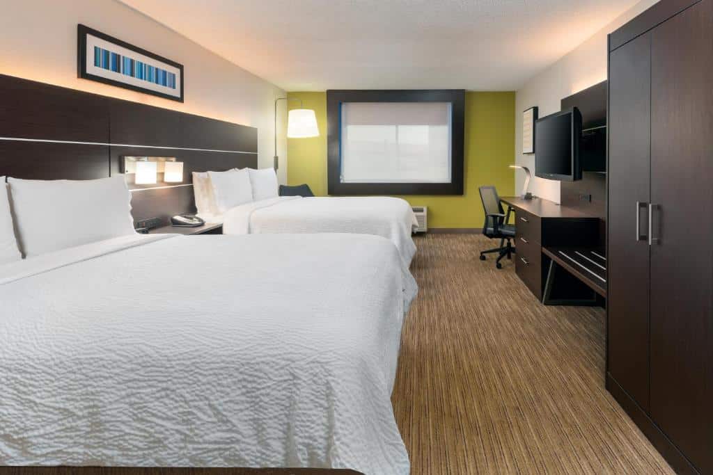 Quarto do Holiday Inn Express Miami Airport Doral Area, an IHG Hotel com duas cama de casal do lado esquerdo da imagem com uma cama de casal a frente no centro uma mesa com telefono e ao fundo uma cama de casal em frente as camas uma mesa de trabalho no centro uma cômoda com TV em cima e um guarda-roupa ao lado, para representar hotéis baratos em Miami.
