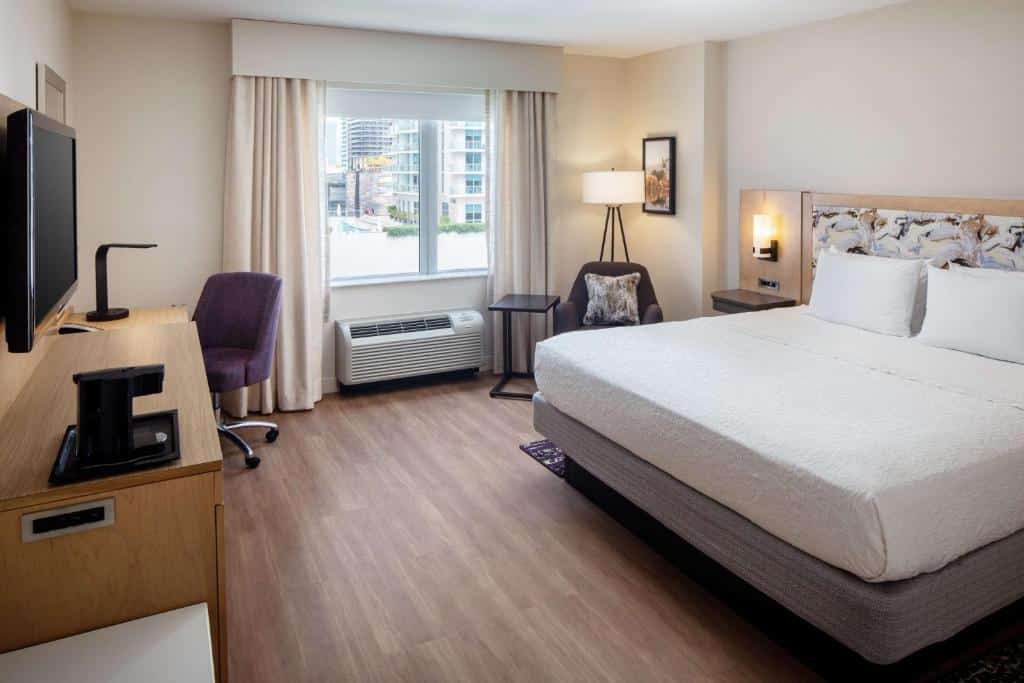 Quarto do Hampton Inn & Suites by Hilton Miami Downtown/Brickell com cama de casal do lado direito da imagem do lado direito uma cadeira com uma mesinha do lado em frente a cama uma cômoda com TV presa na parede, para representar hotéis baratos em Miami.