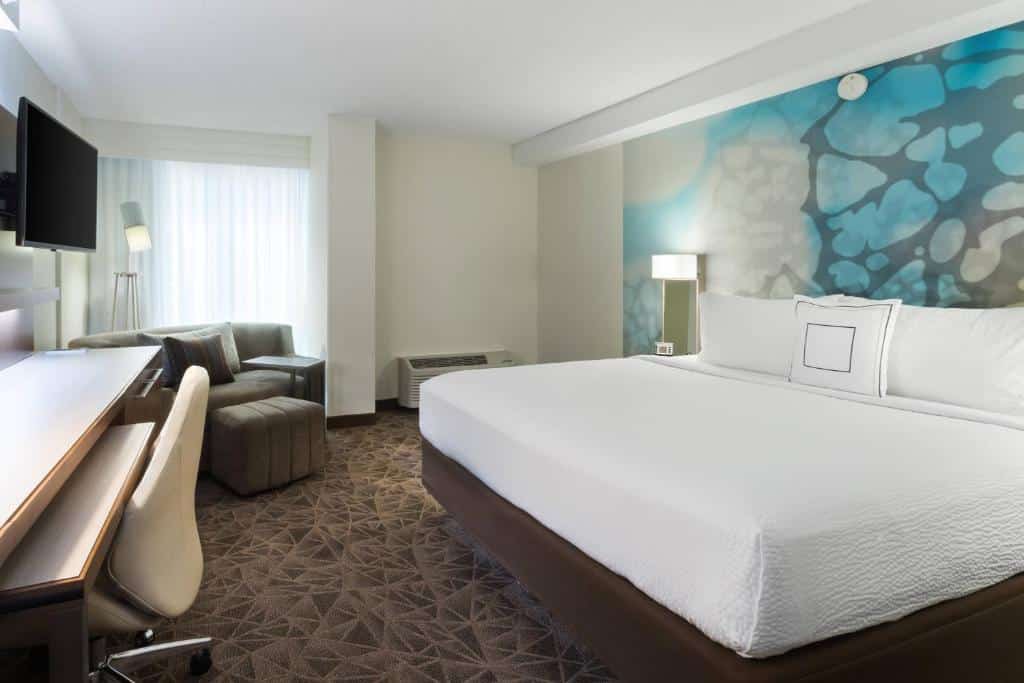 Quarto do Courtyard Miami Downtown Brickell Area com cama de casal do lado direito da imagem, em frente a cama uma cômoda com TV presa na parede do lado direito da cama um pequeno sofá no cama próximo a cômoda, para representar hotéis baratos em Miami.