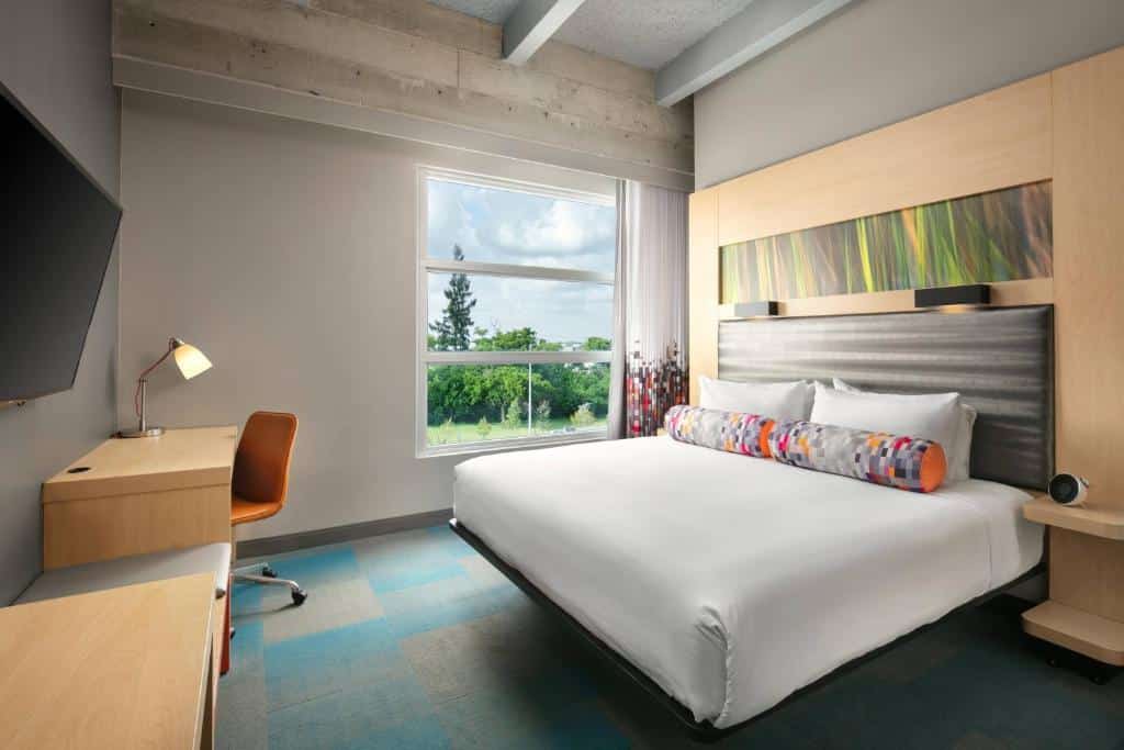 Quarto do Aloft Miami Airport com cama de casal do lado direito da imagem do lado direito da cama uma parede com uma pequena janela em frente a cama uma cômoda com uma cadeira e uma TV presa na parede, para representar hotéis baratos em Miami.