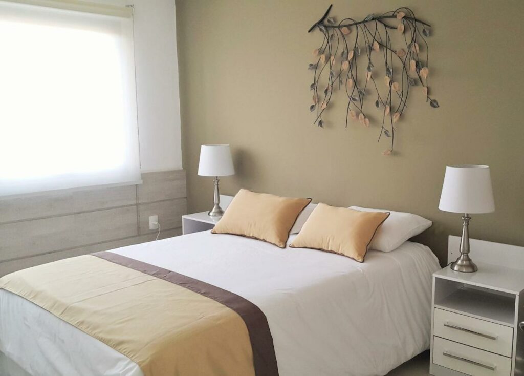 Quarto do Mercosur Universitas. No centro uma cama de casal, de cada lado uma cômoda e um abajur. Do lado esquerdo uma janela com cortina.