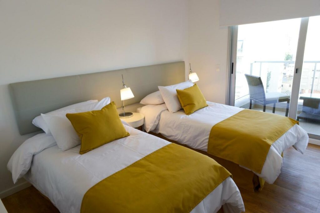 Quarto do Mérit Montevideo Apart & Suites. Duas camas de solteiro e duas estantes com um abajur. No fundo a varanda do quarto.