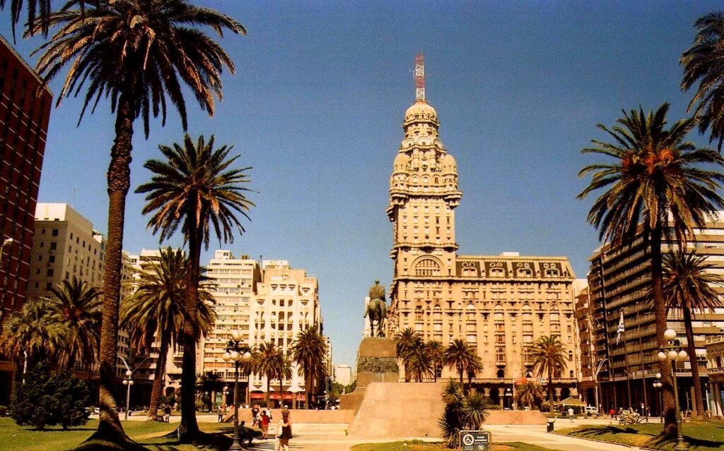 Na frente a Praça da Independência com uma estátua no meio e palmeiras entre os caminhos. No fundo a vista do Palacio Salvo, em Montevidéu.