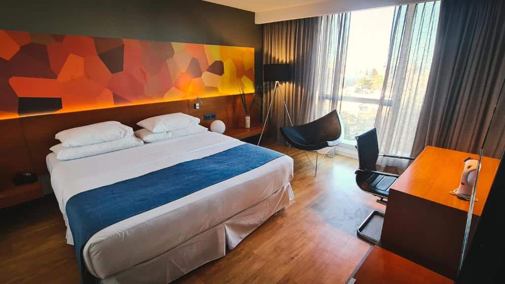 Quarto do BIT Design Hotel. Do lado esquerdo uma cama de casal, um abajur e uma cadeira. No canto direito uma mesa de trabalho, no fundo uma parede de vidro.