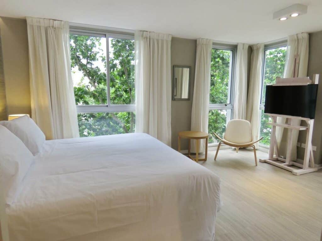 Quarto do Smart Hotel Montevideo. Uma cama de casal do lado esquerdo, do lado direito uma poltrona, uma TV, uma mesa de centro e um espelho. Ao redor paredes de vidro com janelas.