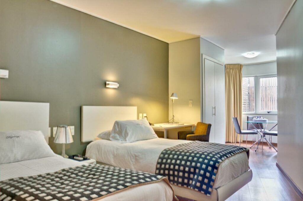 Quarto do Regency Golf - Hotel Urbano. Na frente duas camas de solteiro com abajur entre elas e uma mesa de trabalho, no fundo um guarda-roupa, uma mesa com cadeiras e uma janela.