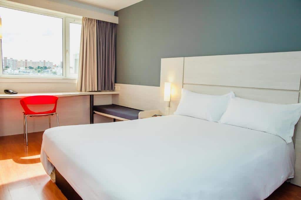 Quarto do ibis Montevideo Rambla. Do lado direito uma cama de casal, no fundo um banco e uma mesa de trabalho, no fundo a janela do quarto.