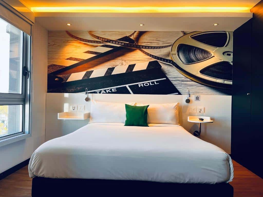Quarto do ibis Styles Montevideo Biarritz. No centro a cama de casal, de cada lado uma estante com uma luminária. Do lado direito um guarda-roupa, do lado esquerdo uma janela.