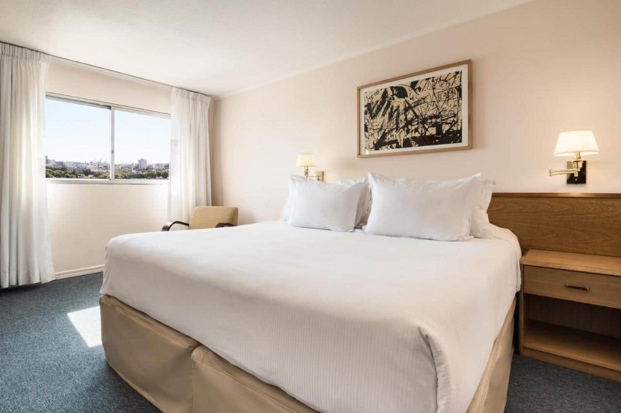 Quarto do Days Inn by Wyndham Montevideo. Uma cama de casal no centro, de cada lado uma cômoda e um lustre. No fundo uma poltrona e uma janela.