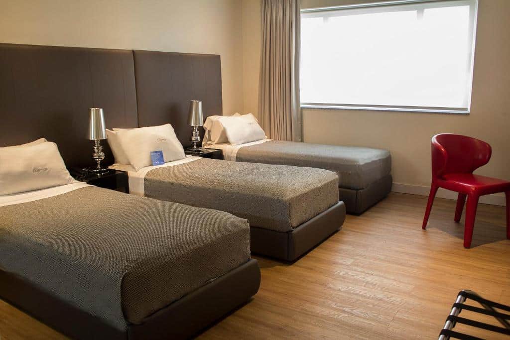 Quarto do Regency Way Montevideo Hotel, com três camas de solteiro do lado esquerdo, entre elas uma cômoda e um abajur. Do lado direito uma cadeira, no fundo uma janela.
