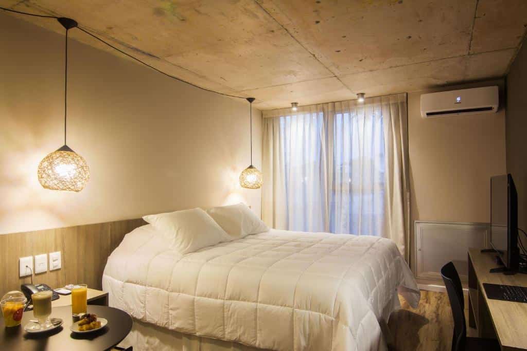 Quarto do Hotel Ciudadano Suites. Uma cama de casal no centro com um lustre de cada lado. Do lado esquerdo uma mesa com comida, do lado direito uma mesa de trabalho com TV. No fundo uma janela com cortina e um ar-condicionado.