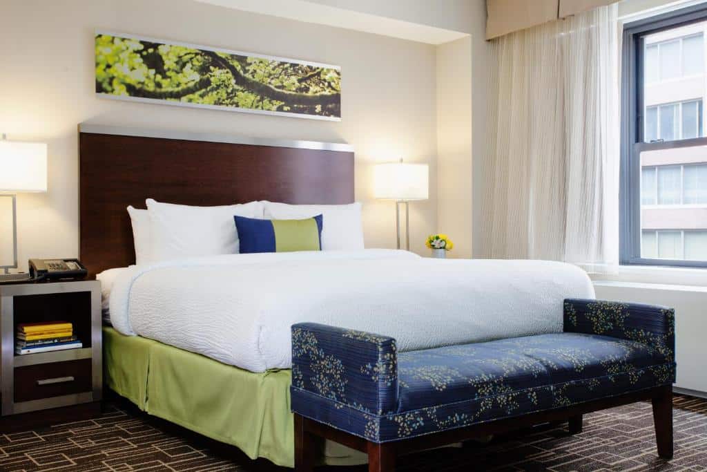 Um quarto do Residence Inn by Marriott hotel com uma cama bem arrumada, dois abajures, um banco azul ao pé e uma janela que deixava entrar a luz do dia.