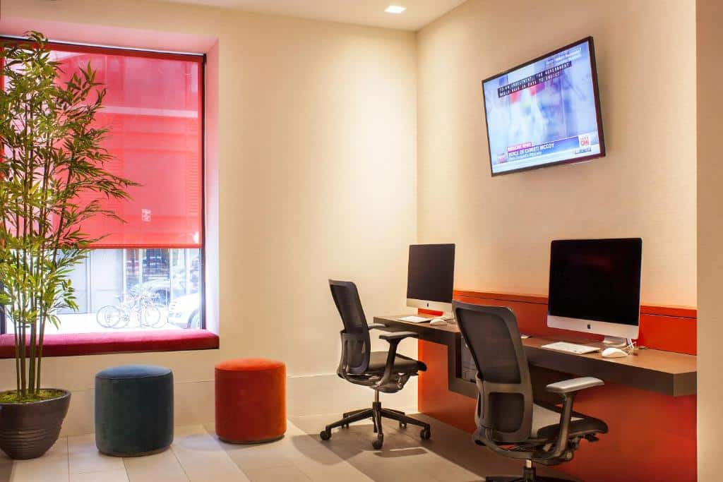 Área de escritório moderna do Hotel 46 com dois computadores de mesa, duas cadeiras, uma TV na parede, um vaso de plantas e dois pufes redondos. Este é um dos hotéis baratos em Nova York.