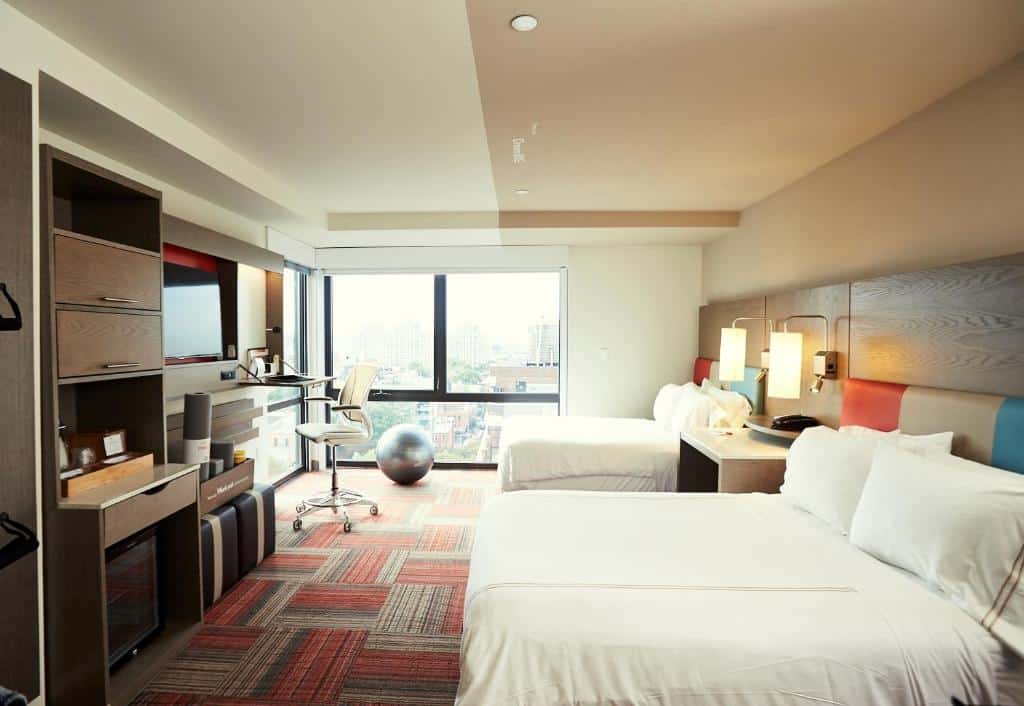 quarto do EVEN Hotel Brooklyn com duas camas, uma escrivaninha com cadeira, bola de ginástica, janela grande e unidades de armazenamento embutidas.