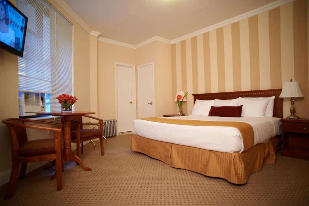 quarto do Hotel St. James com uma cama king-size, parede com detalhes listrados, mesa pequena com cadeiras e uma TV na parede.