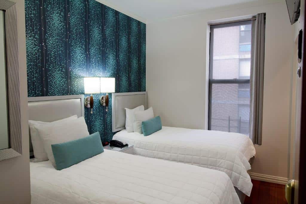quarto do Belnord Hotel com duas camas de solteiro, travesseiros com detalhes em azul-petróleo, papel de parede estampado e uma janela com vista para a cidade.