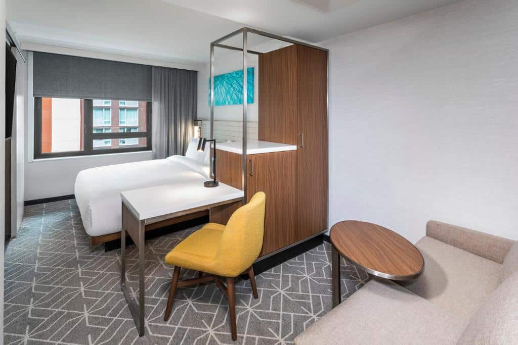 quarto do SpringHill Suites by Marriott com uma cama, escrivaninha e cadeira de madeira, sofá, mesa redonda e janela grande com persianas cinza fechadas.
