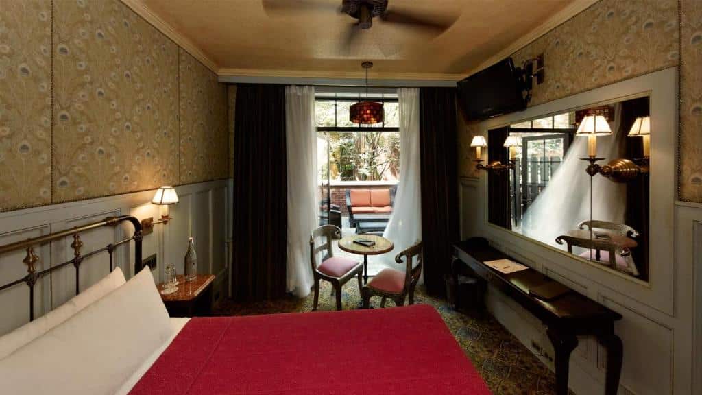 quarto do The Jane Hotel com uma cama, mesa e cadeiras pequenas, espelho de parede, TV e uma varanda com assentos ao ar livre visível através de portas de vidro.