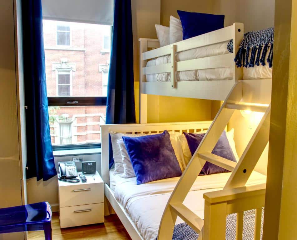 Pequeno quarto do The Ridge com um beliche, roupa de cama branca, travesseiros azuis, uma janela e uma mesa de cabeceira com um telefone e um bloco de anotações.