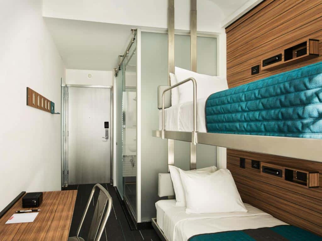 quarto do Pod Times Square, bem compacto, com beliches, uma mesa e cadeira de madeira e um banheiro com paredes de vidro perto da entrada.