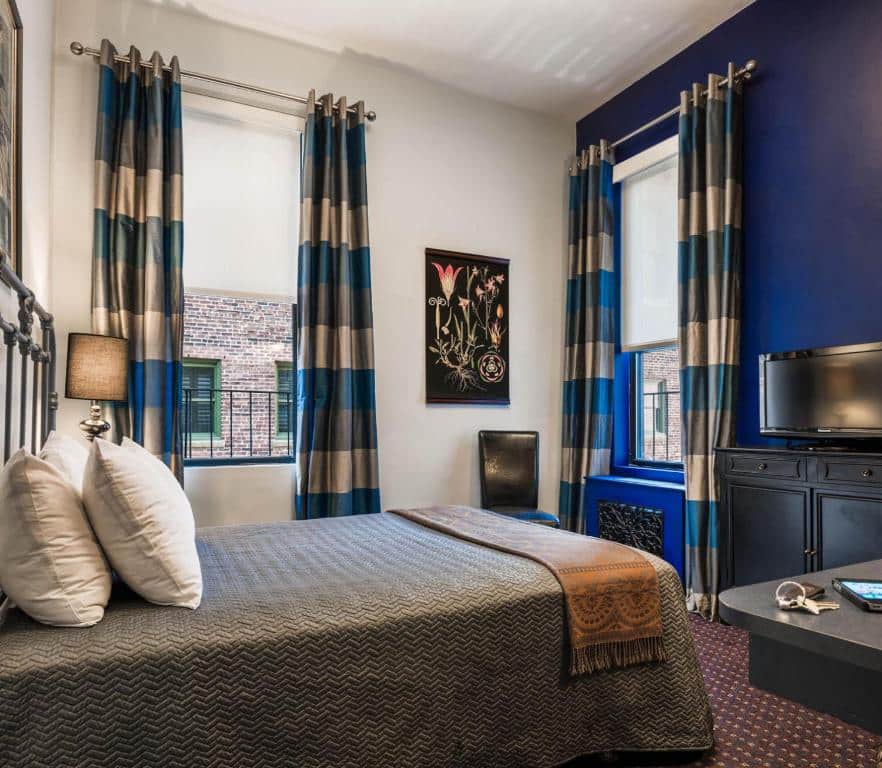 Quarto do Seton Hotel com uma cama bem arrumada, paredes azuis, cortinas listradas, uma TV e uma janela que mostrava um prédio de tijolos do lado de fora.