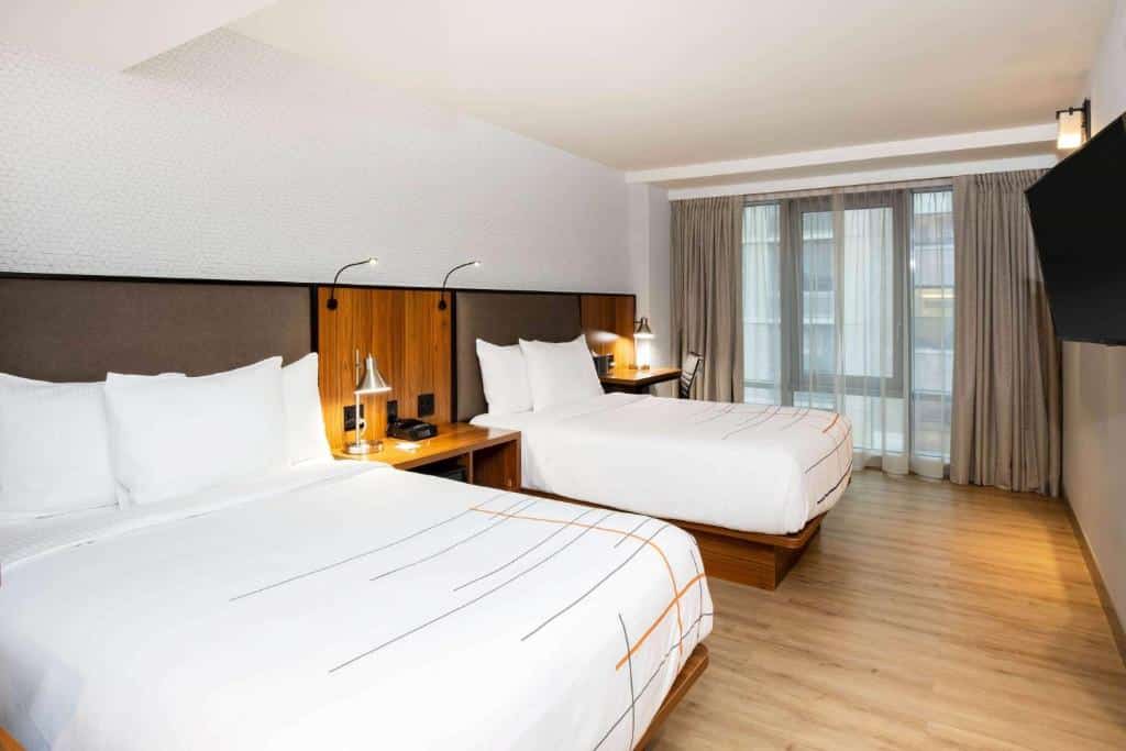 Quarto do hotel La Quinta by Wyndham com duas camas de casal, decoração moderna, janela grande com cortinas, piso de madeira e uma TV na parede.