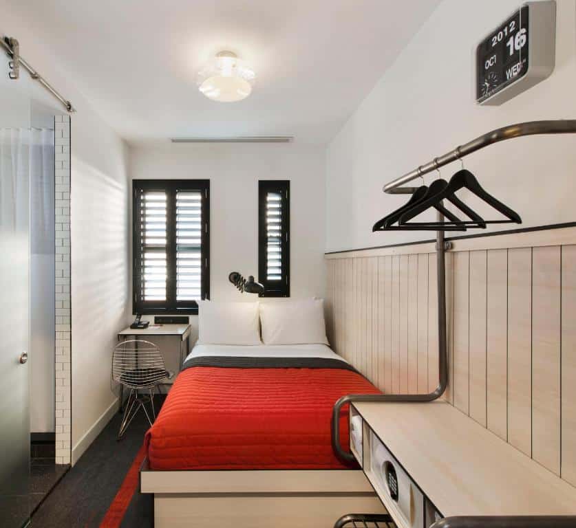 quarto do Pod 39, pequeno e moderno, com uma cama de casal, cobertor vermelho, escrivaninha, cadeira, armário aberto e um relógio de parede.