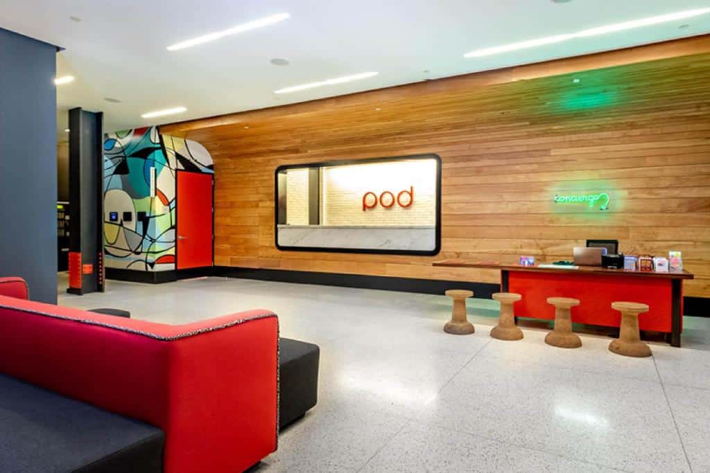 Saguão do hotel moderno Pod 39 com assentos vermelhos, uma parede de madeira com a inscrição "pod" e arte abstrata colorida perto de uma porta vermelha. É um dos hotéis baratos em Nova York.