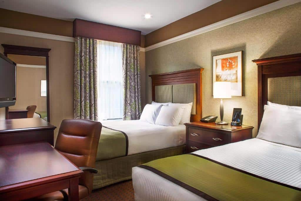 quarto do Hotel 46 com duas camas de casal, uma escrivaninha e cadeira, quadro na parede, luminária, janela com cortinas e uma TV.