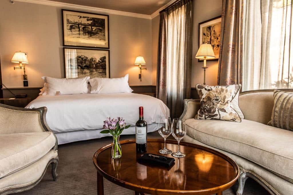 Quarto do The Singular Santiago com cama de casal ao fundo a frente uma mesa redonda com uma garrafa de vinho e duas taças e sofá ao lado da mesa.