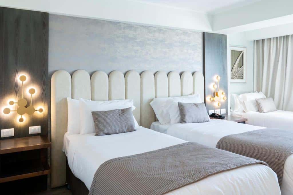 Um quarto do Hotel He Wang Santiago com duas camas de casal, lençóis brancos, mantas cinza, luminárias de parede e uma mesa de cabeceira entre as camas.