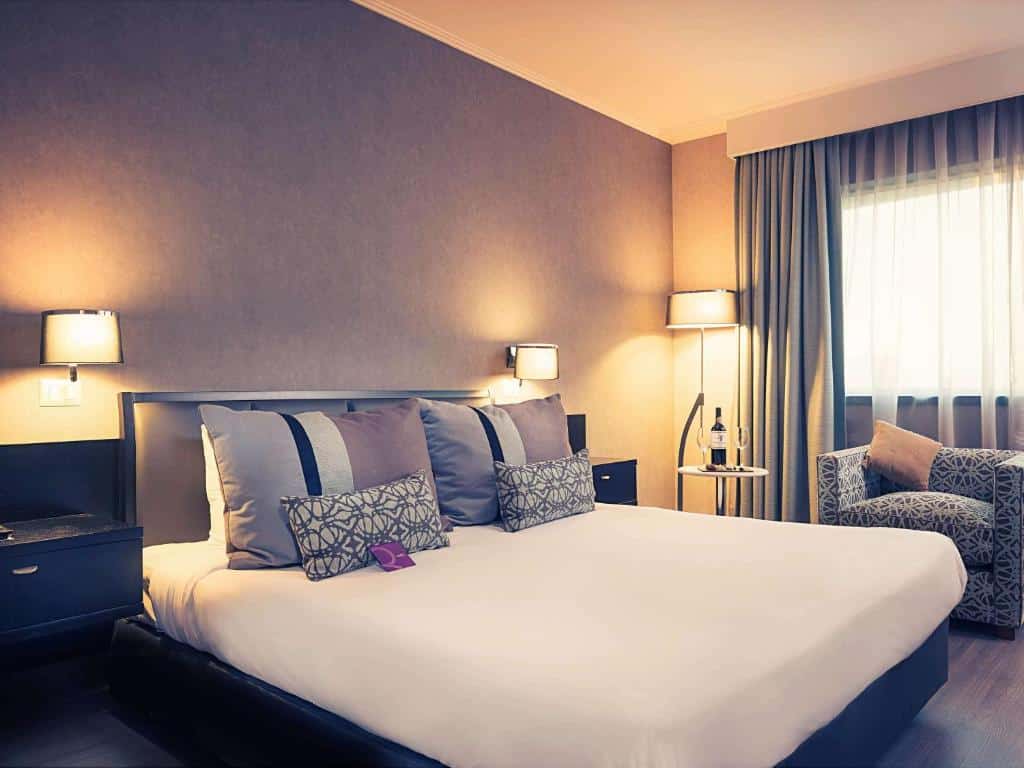Quarto do Mercure Santiago Centro com cama de casal no centro do quarto duas cômodas ao lado da cama e uma poltrona do lado esquerdo.