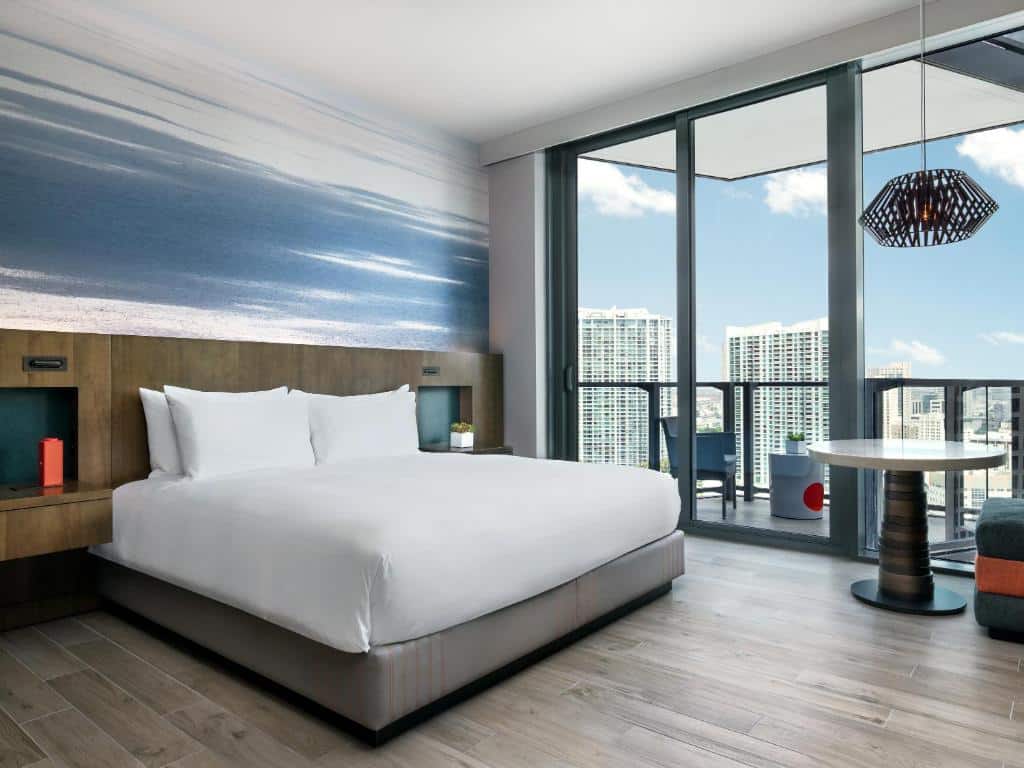 Quarto do East Miami com cama de casal, duas cômodas ao lado da cama, uma mesa-redonda do lado esquerdo e portas de vidro que dá acesso a varanda.