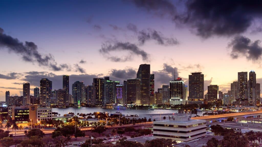 Vista de Port Miami Tunnel, Miami no final do dia com prédios enormes com luzes acessa e um lago em frente.