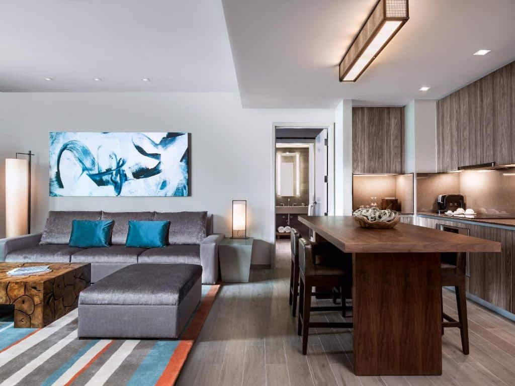 Sala de estar e cozinha comacta do East Miami com sofá cinza com almofadas azul turquesa no lado esquerdo com mesa de madeira ao centro. Do lado direito mesa de três lugares com armários ao fundo, para representar hotéis com cozinha em Miami.