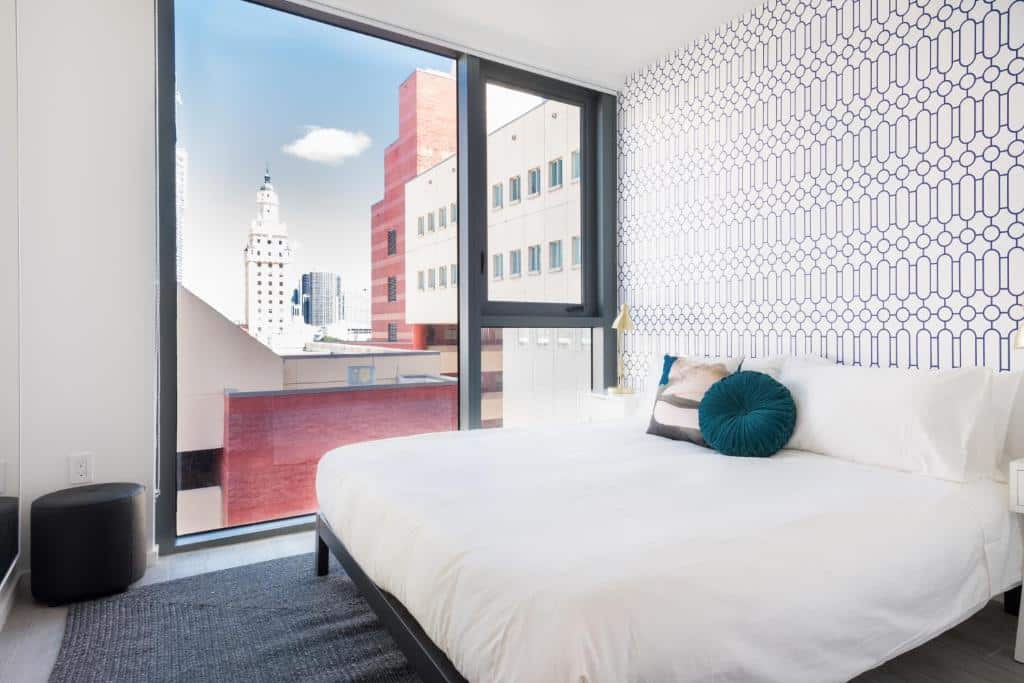 Quarto moderno no The Guild Downtown | X Miami com janela grande, parede com detalhes estampados, roupa de cama branca e vista para os prédios da cidade do lado de fora, para representar hotéis com cozinha em Miami.