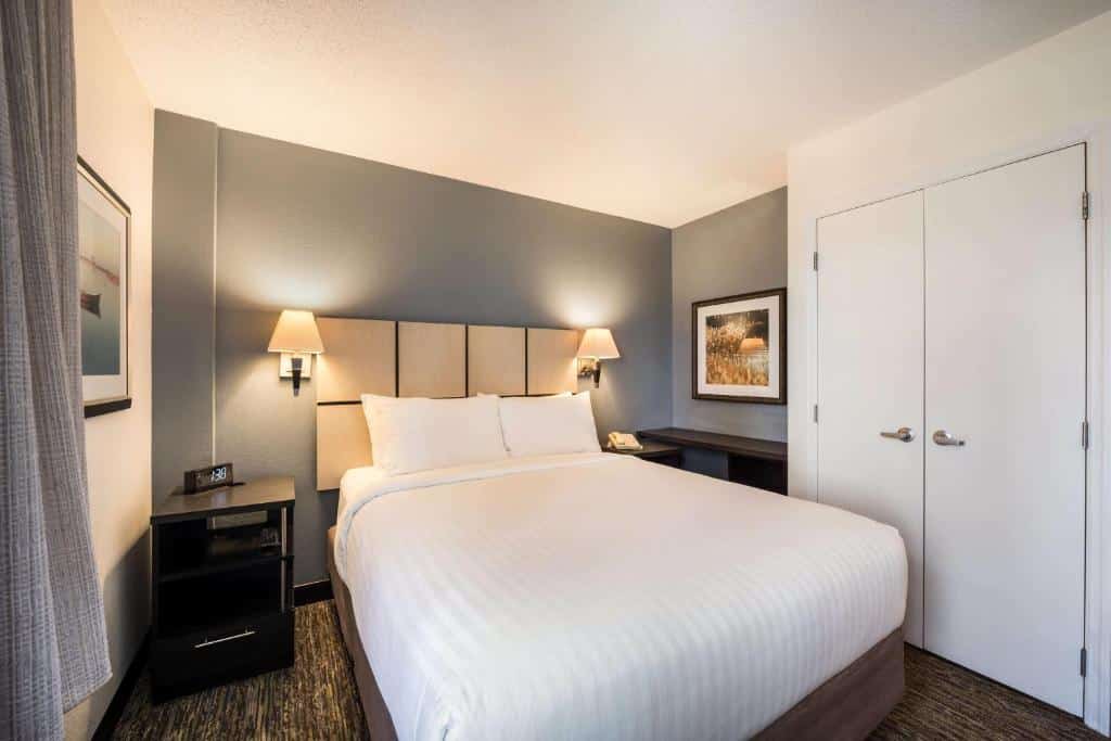 Um quarto no Sonesta Simply Suites Miami Airport Doral com uma cama bem arrumada, dois abajures, quadros emoldurados, uma mesa de cabeceira e uma porta dupla fechada, para representar hotéis com cozinha em Miami.
